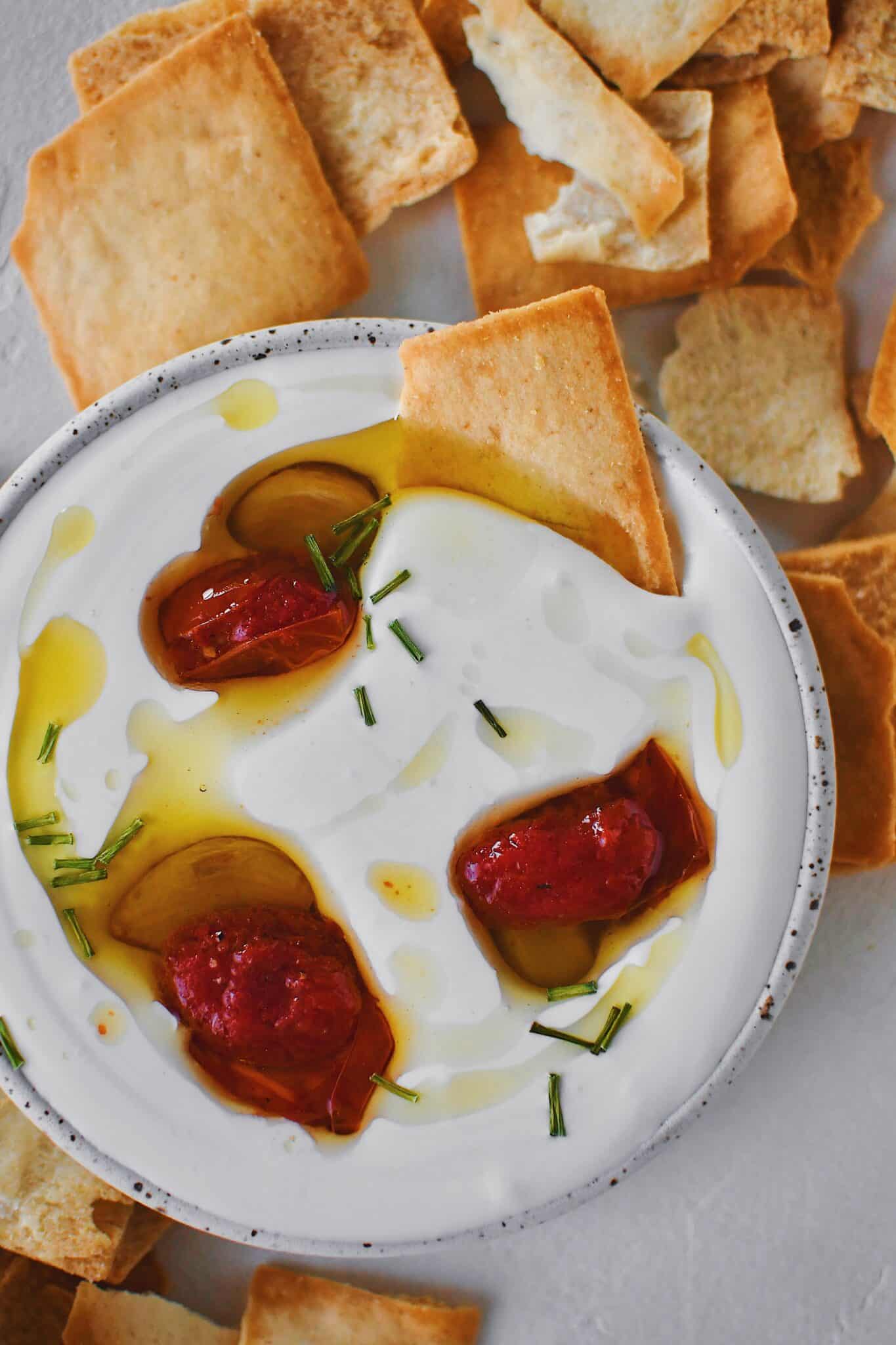A pita chip dipped in whipped feta dip that has been served with confit garlic and tomatoes with chives on top.