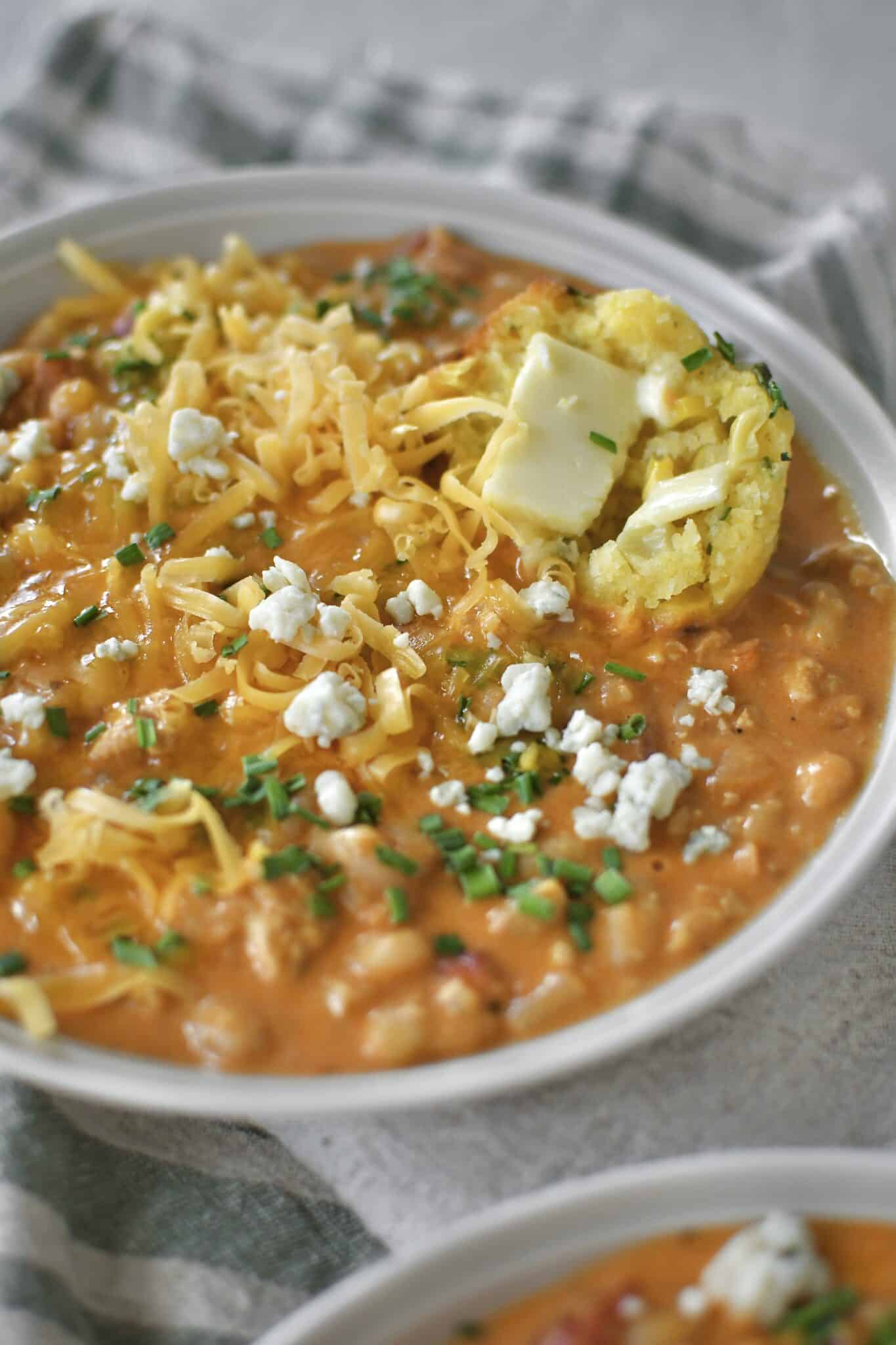 Buffalo Chicken Chili served in a bowl, topped with chives, blue cheese, and cheddar cheese. And a piece of cornbread muffin dunked in it.