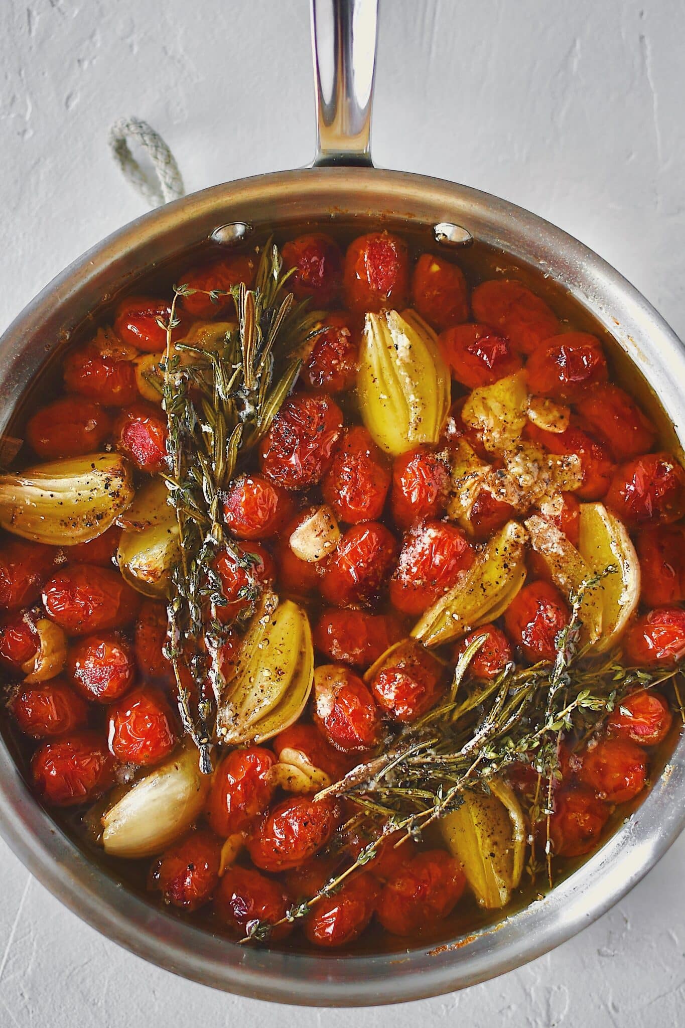 Grape tomatoes in a pan with shallots, sliced garlic, thyme, rosemary, salt, and pepper after roasting and made into confit.