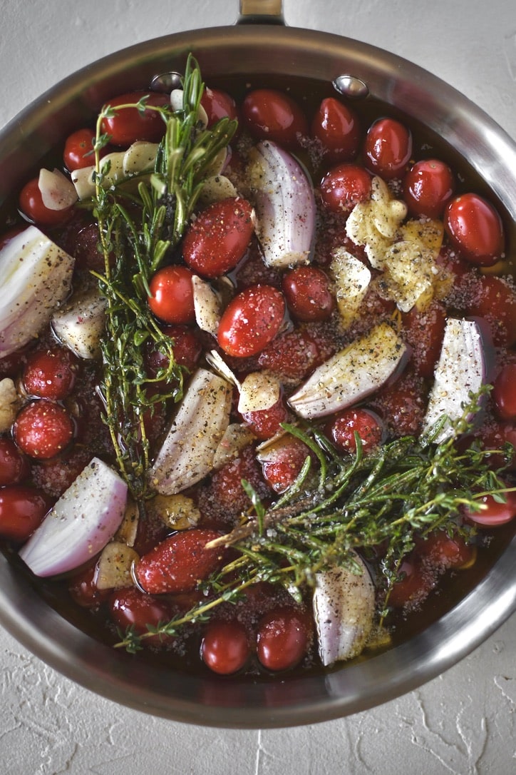 Grape tomatoes in a pan with shallots, sliced garlic, thyme, rosemary, salt, and pepper ready to be roasted and made into confit.