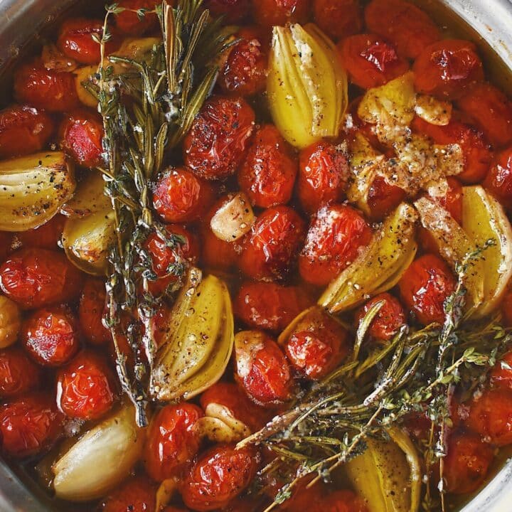 Grape tomatoes in a pan with shallots, sliced garlic, thyme, rosemary, salt, and pepper after roasting and made into confit.