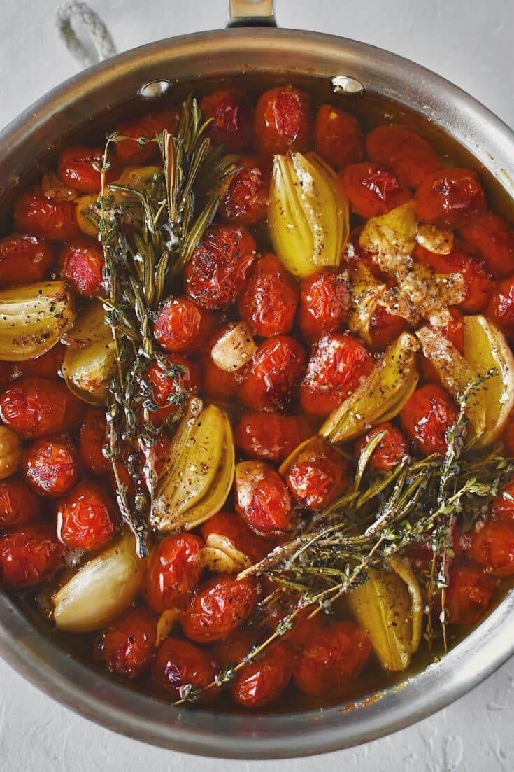 Grape tomatoes in a pan with shallots, sliced garlic, thyme, rosemary, salt, and pepper after roasting and made into confit.