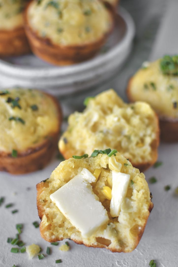 Cornbread Muffins scattered on counter, one split in half with butter on it.