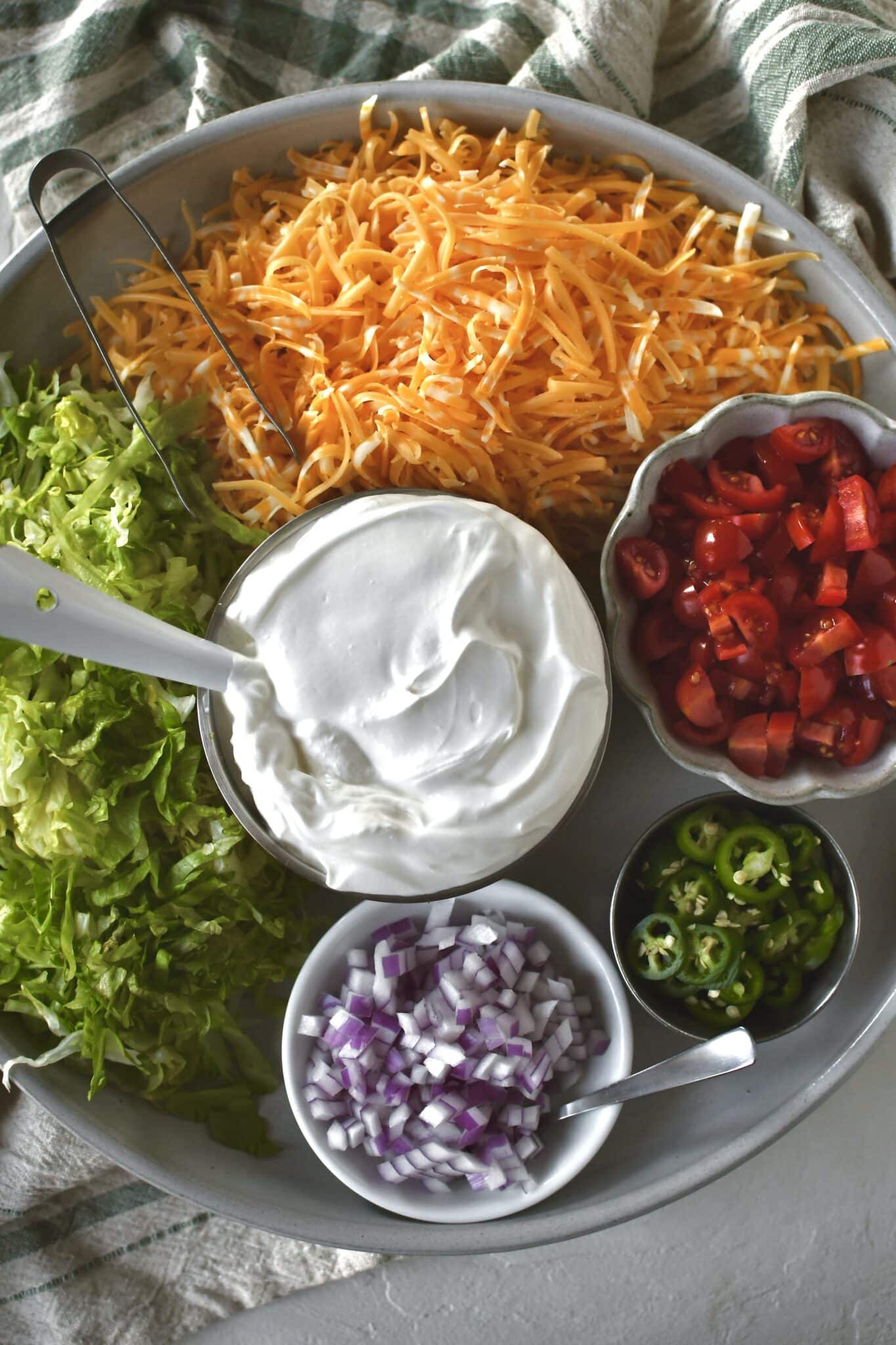 Optional toppings for tacos on a platter. Cheese, shaved lettuce, diced tomatoes and onions, sour cream, and sliced spicy peppers.