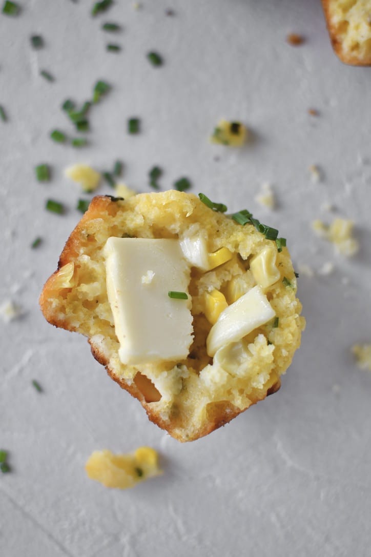 Half a Cornbread Muffin with crumbs all around it on a counter and butter on the muffin.