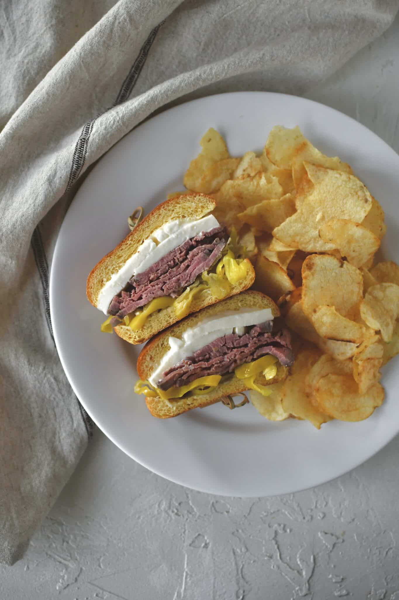 Fully assembled roast beef sandwich, cut in half and on a plate with some chips ready to eat.