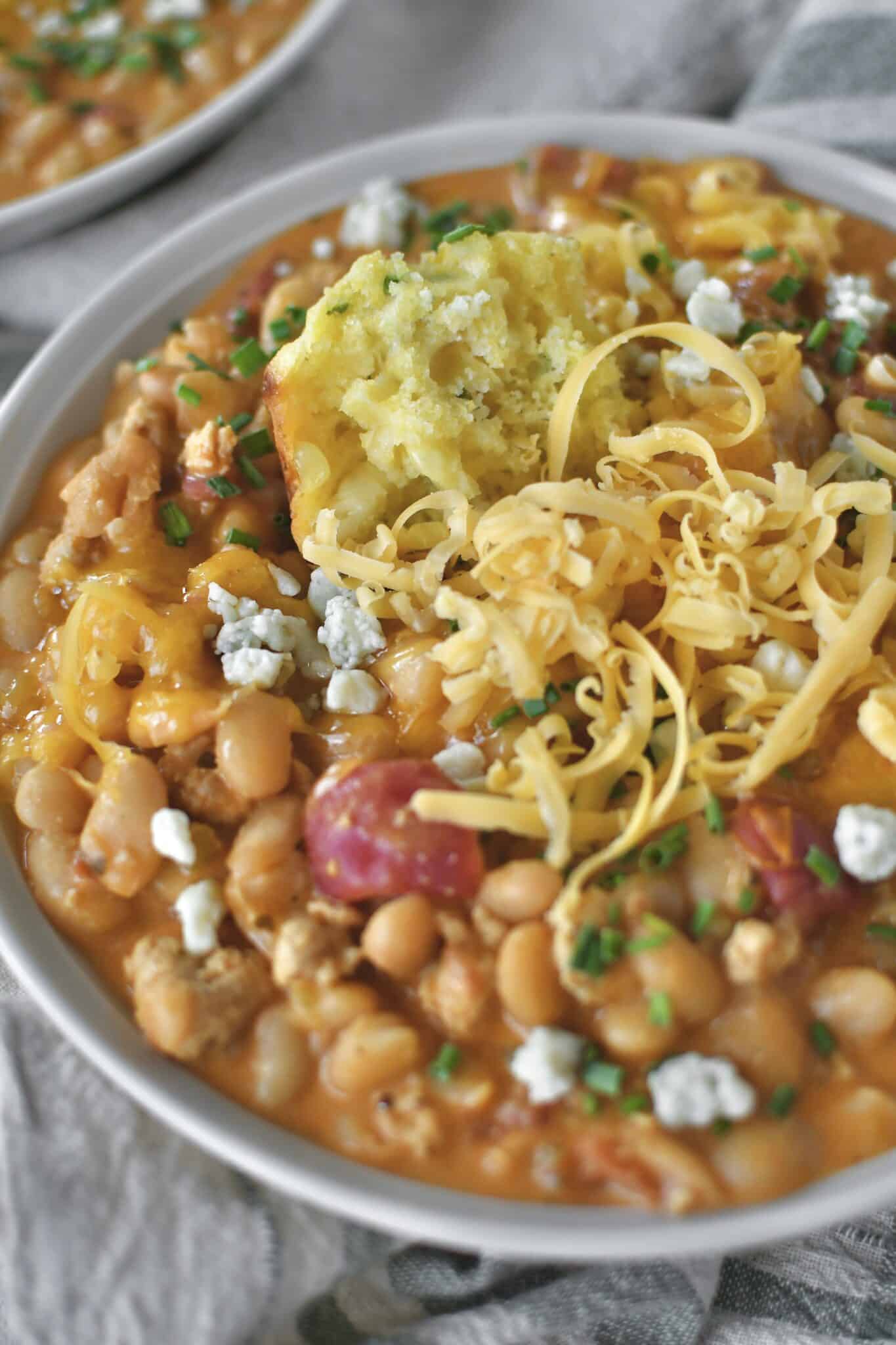 Buffalo Chicken Chili served in a bowl, topped with chives, blue cheese, and cheddar cheese. And a piece of cornbread muffin dunked in it.