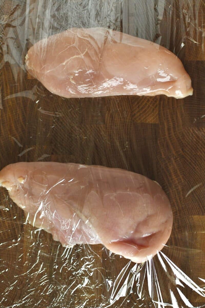 Chicken breasts on a cutting board between two pieces of cling film, before tenderizing to flatten.