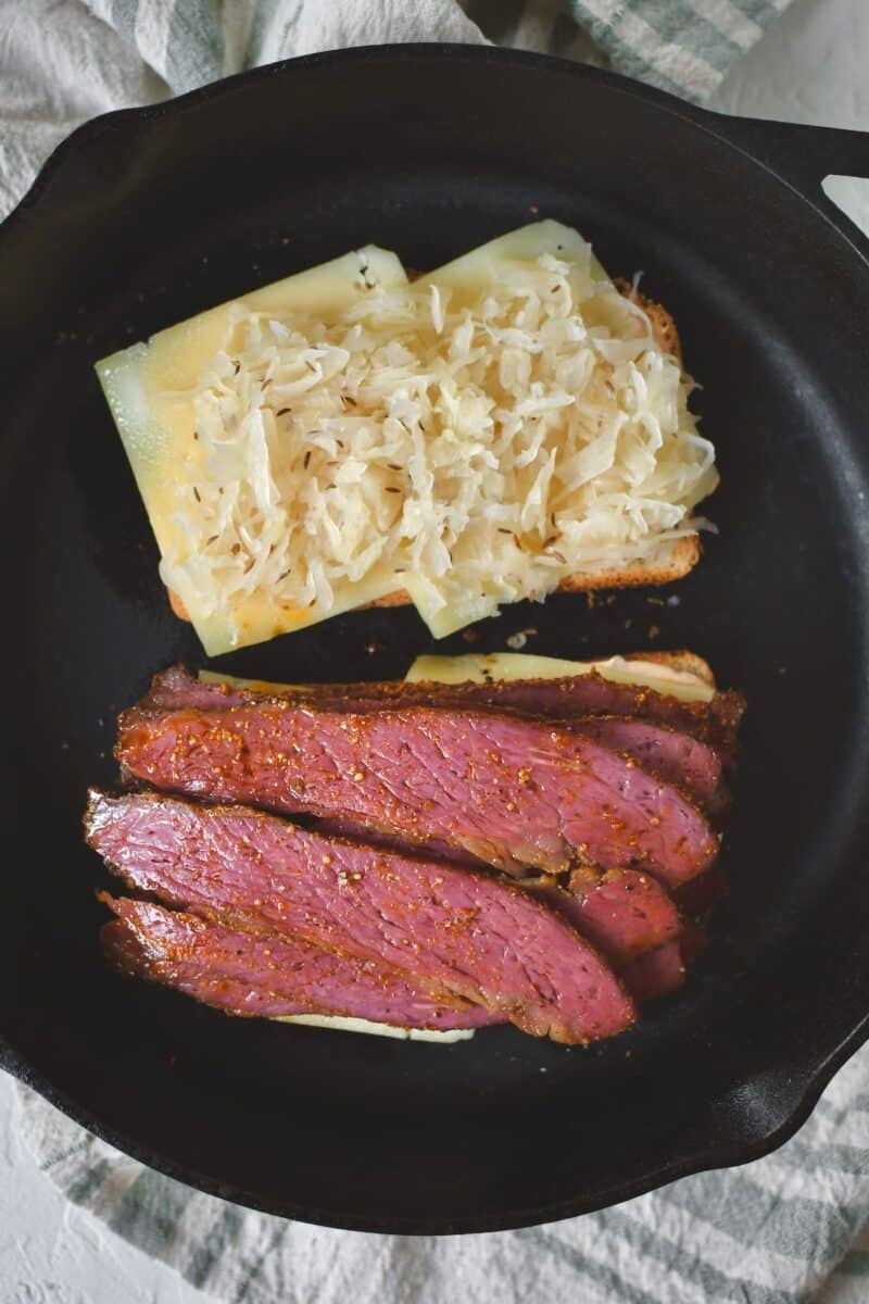 Placing the steamed pastrami and some sauerkraut on the bread before assembling the sandwich.