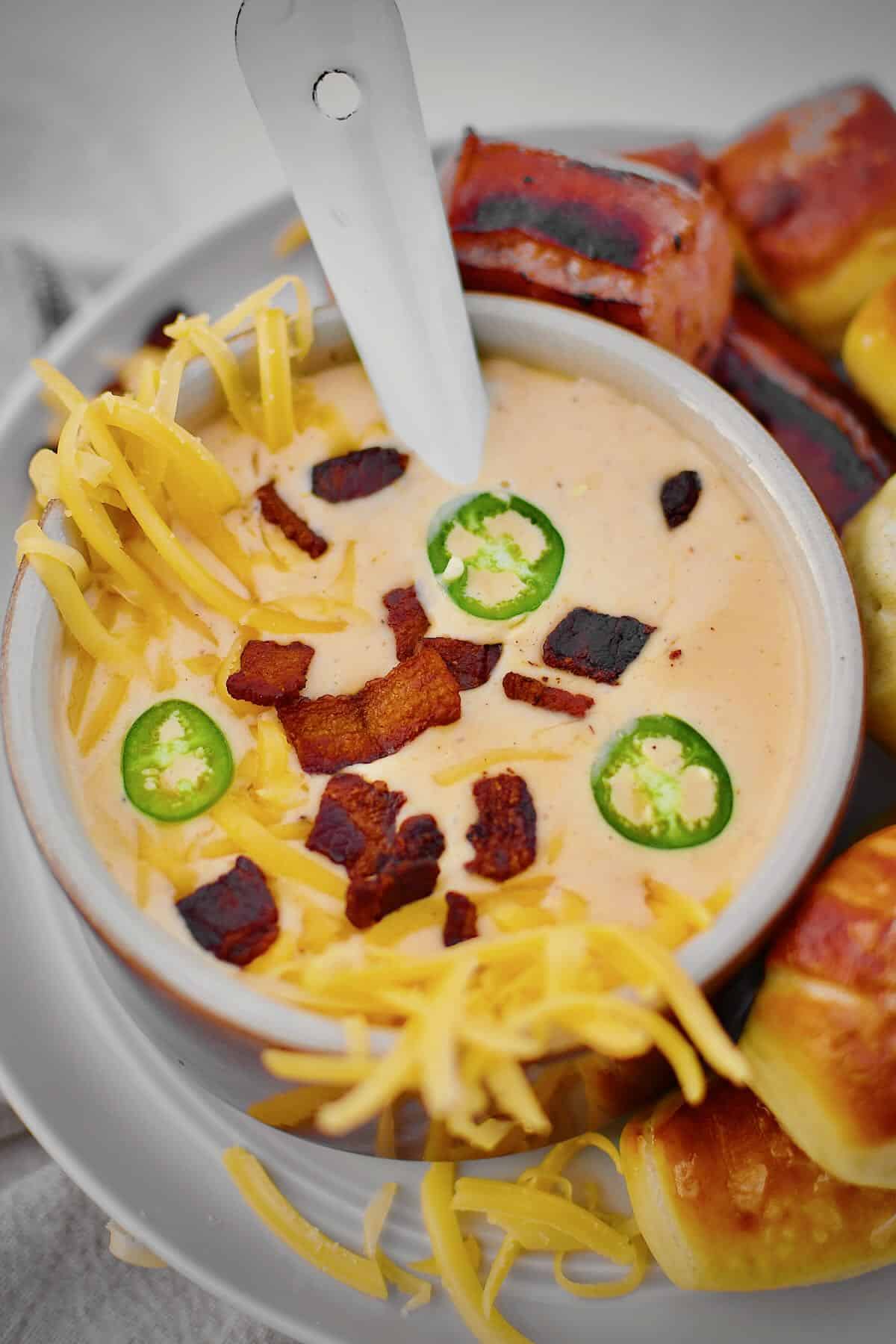 Beer Cheese Soup topped with more cheese, bacon bits, and serrano chili slices, ready to eat. Served with Kielbasa and Pretzel Bites