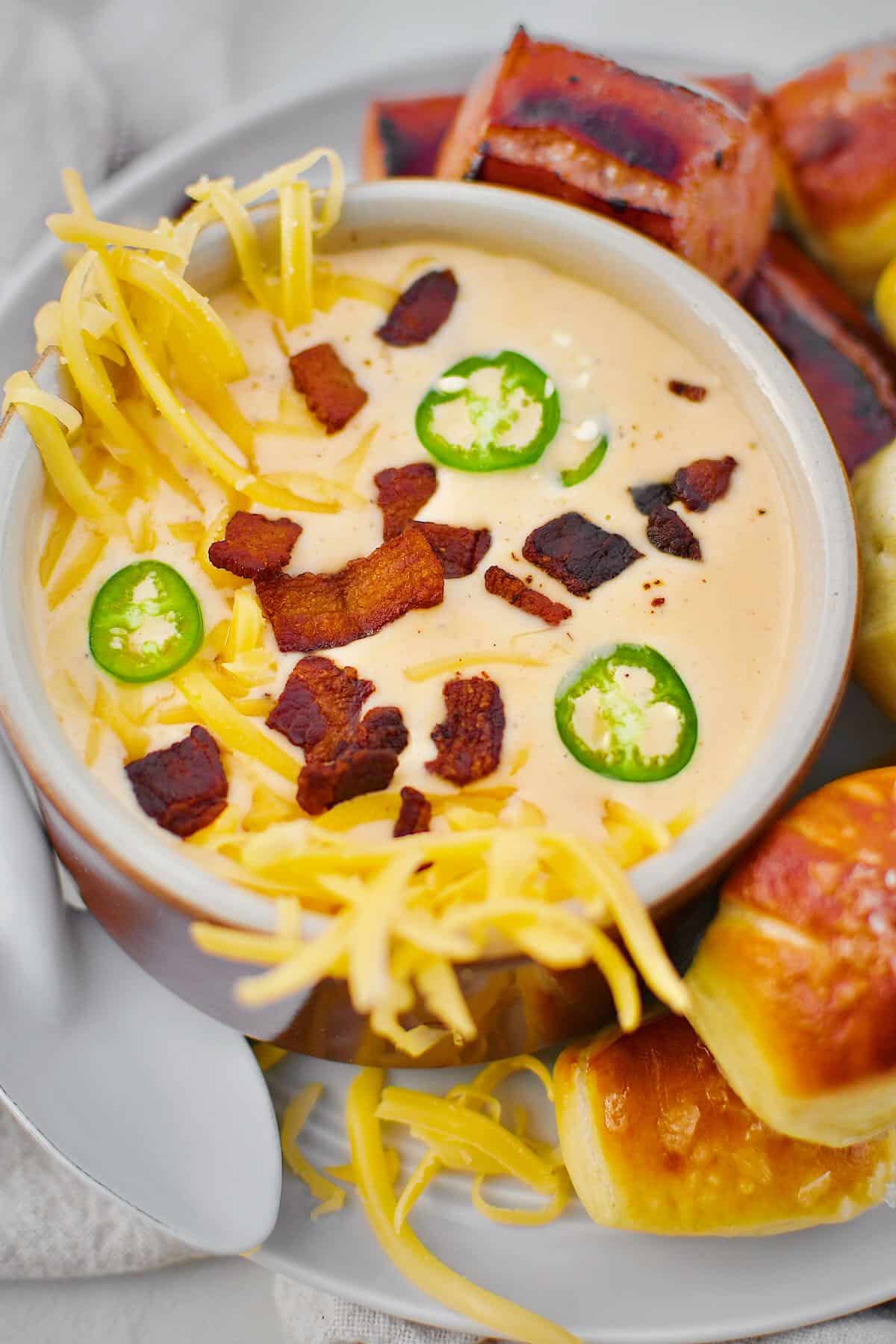 Beer Cheese Soup topped with more cheese, bacon bits, and serrano chili slices, ready to eat. Served with Kielbasa and Pretzel Bites