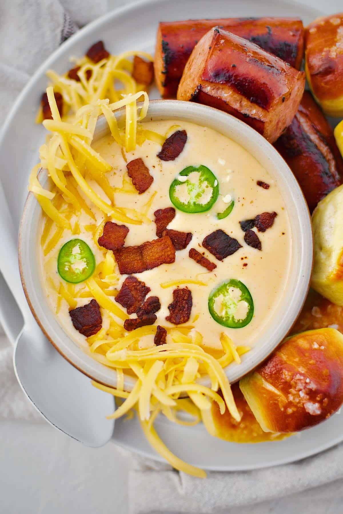 Beer Cheese Soup topped with more cheese, bacon bits, and serrano chili slices, ready to eat. Served with Kielbasa and Pretzel Bites