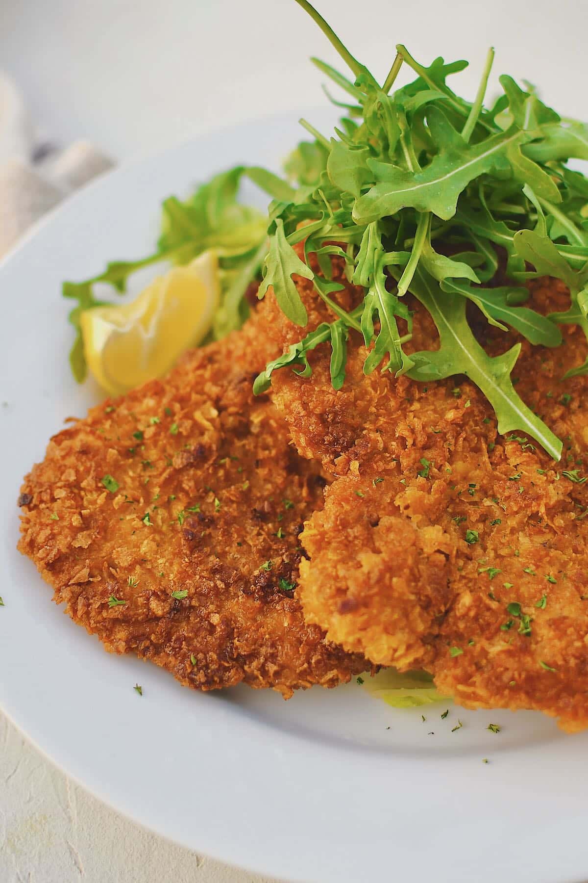 Pork Schnitzel on a plate, served with an arugula salad and lemon wedges.