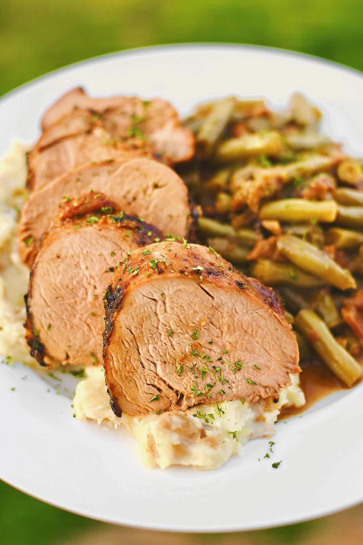 Slices of pork tenderloin on a plate with mashed potatoes under it and southern green beans on the side.