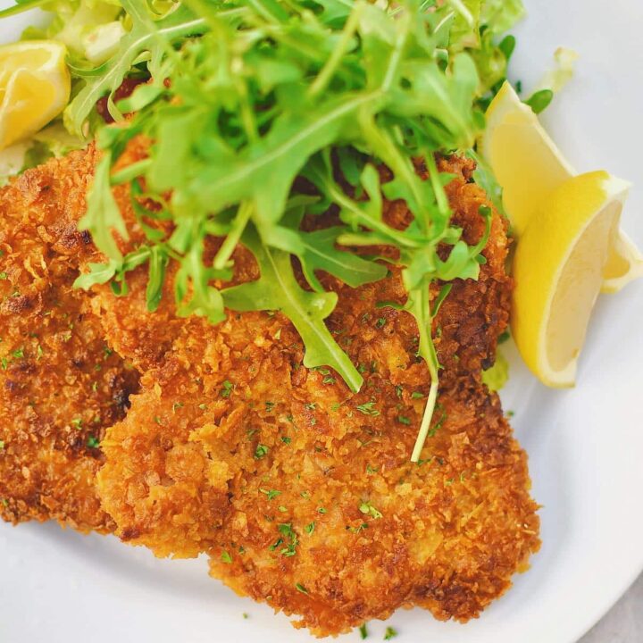 Pork Schnitzel on a plate, served with an arugula salad and lemon wedges.