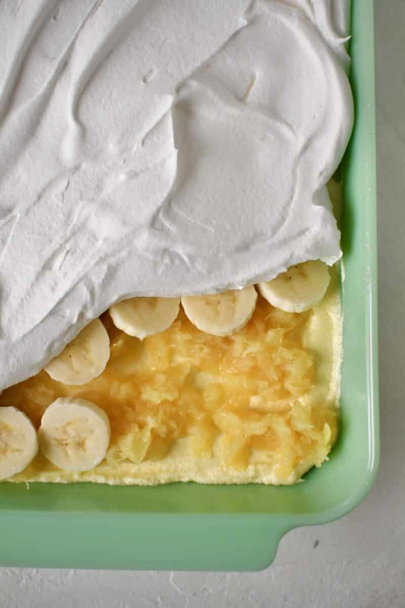 Finishing the cake by spreading out the crushed pineapple over the filling, adding a layer of bananas and finishing with the whipped topping.