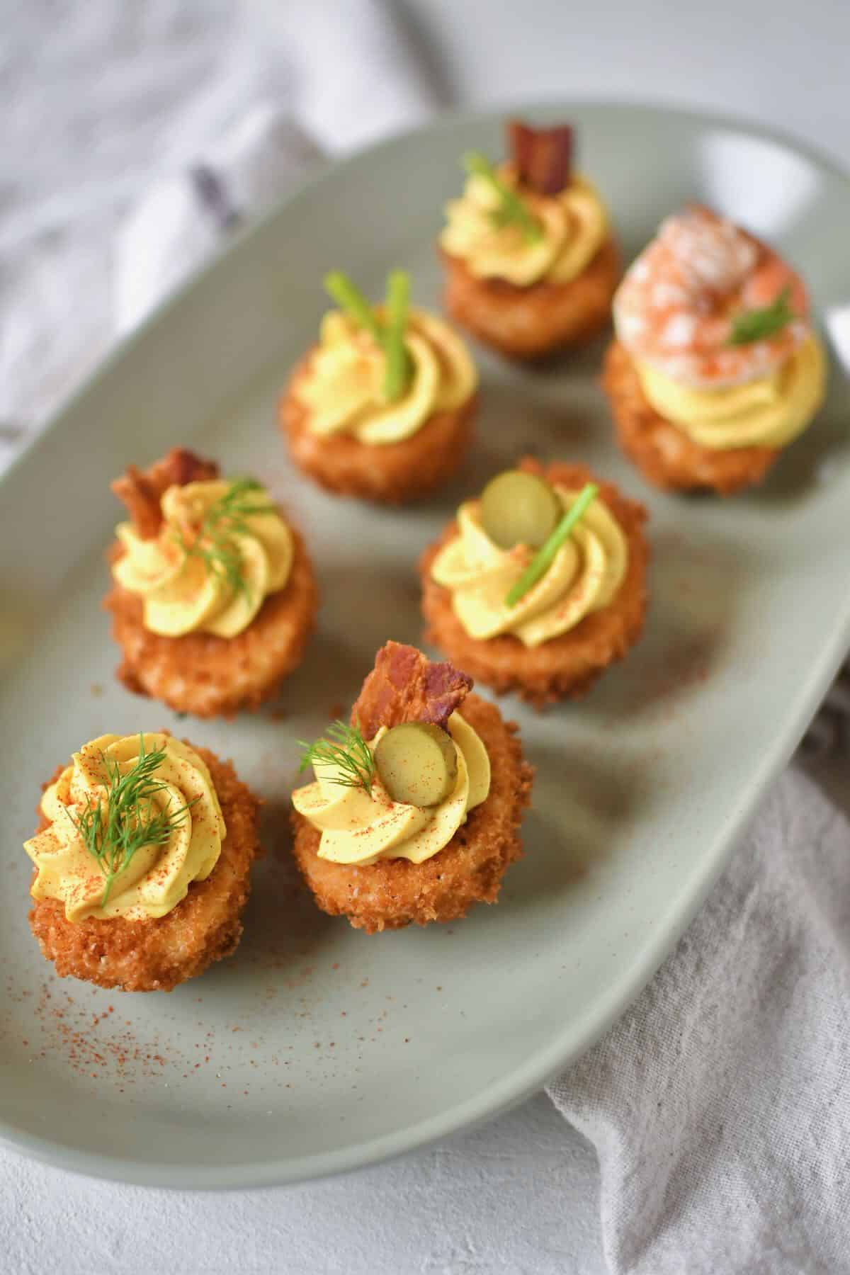 Finished Deep Fried Deviled Eggs on a platter topped with an array of optional toppings like sliced gherkins, herbs, bacon, and shrimp.