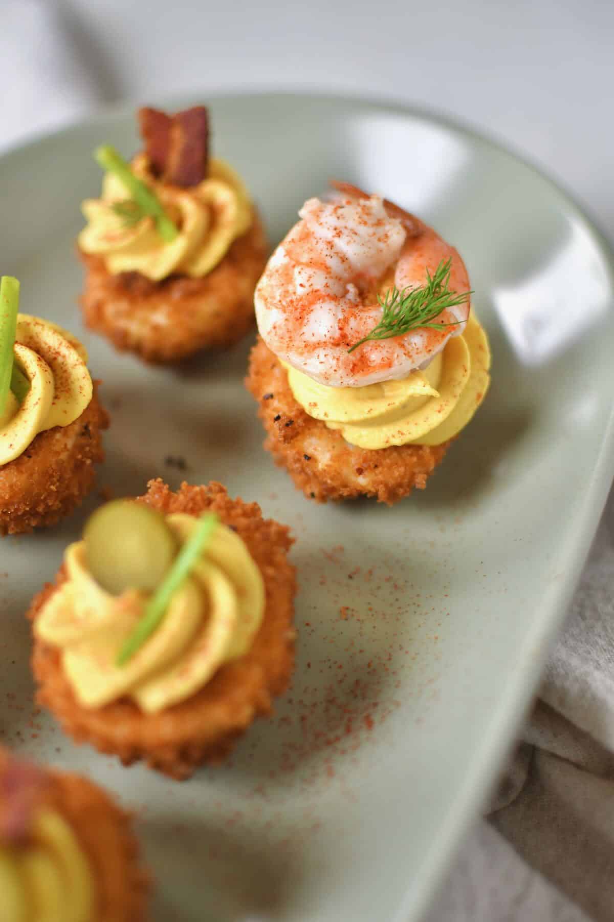 Finished Deep Fried Deviled Eggs on a platter topped with an array of optional toppings like sliced gherkins, herbs, bacon, and shrimp.