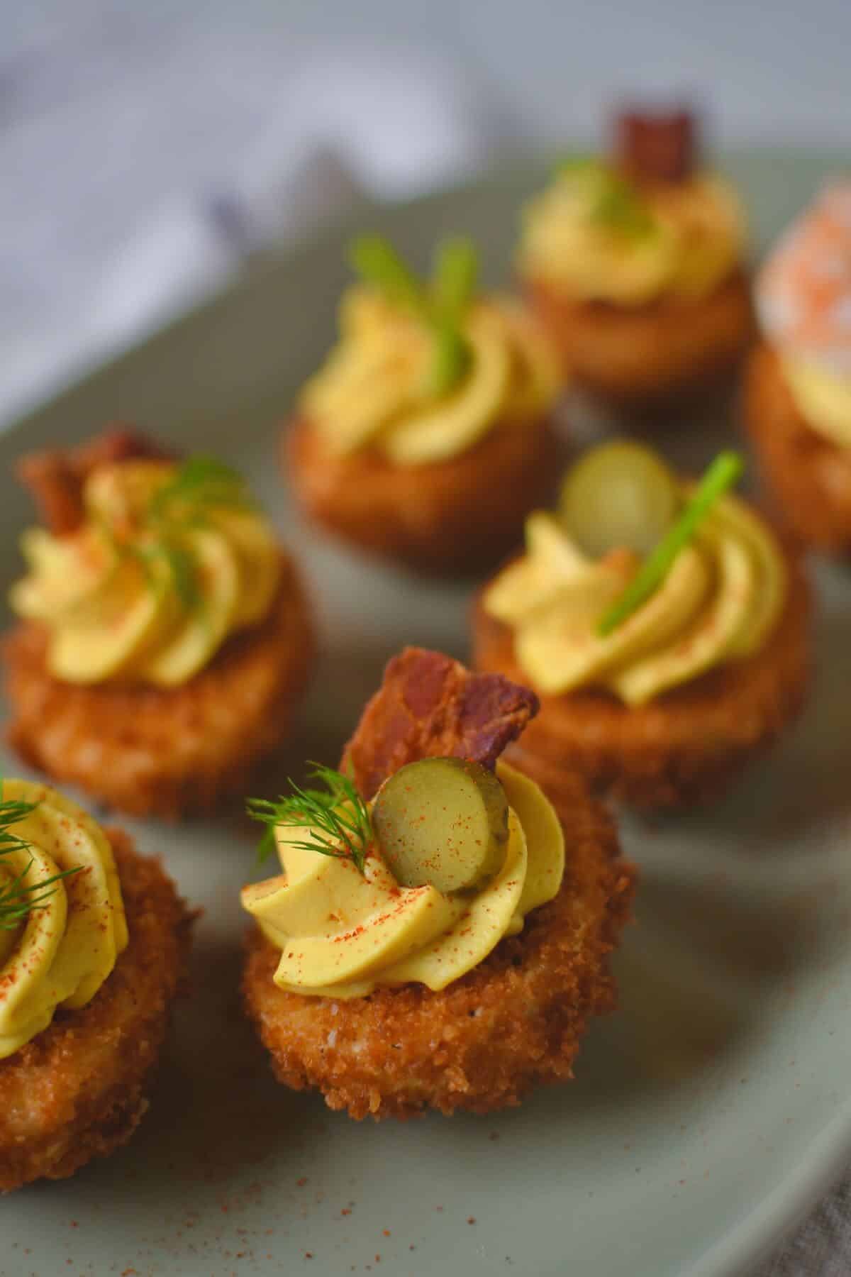 Finished Deep Fried Deviled Eggs on a platter topped with an array of optional toppings like sliced gherkins, herbs, bacon, and shrimp.