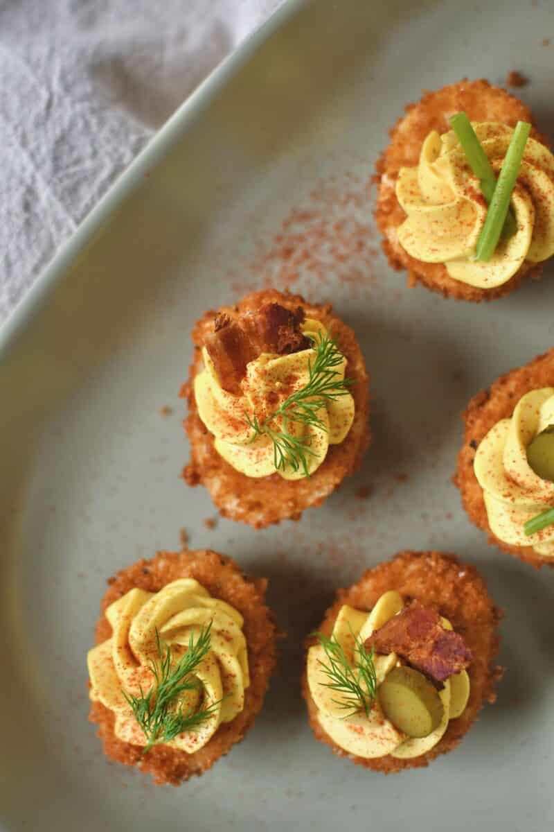 Finished Deep Fried Deviled Eggs on a platter topped with an array of optional toppings like sliced gherkins, herbs, bacon, and shrimp.