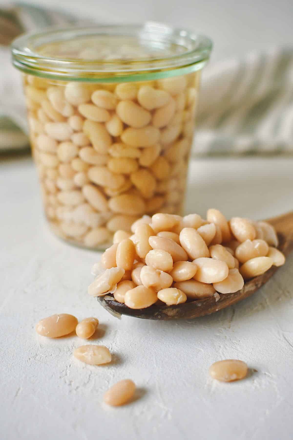 Cooked beans, stored in a glass jar for later use. some laying in a spoon on the table on the side of the jar.