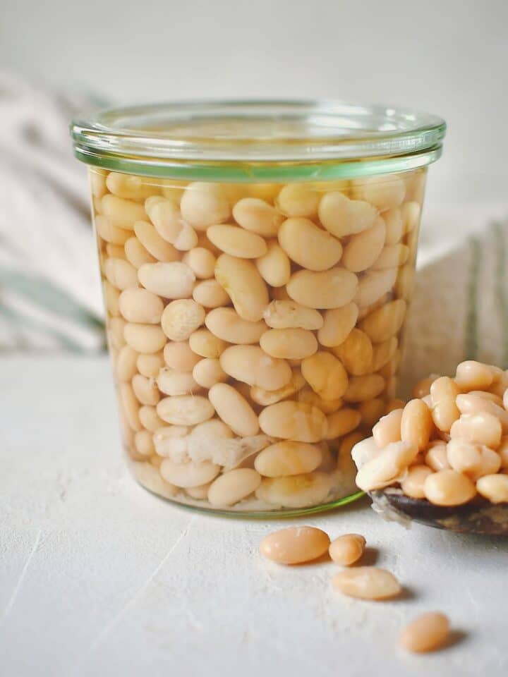 Cooked beans, stored in a glass jar for later use. some laying in a spoon on the table on the side of the jar.