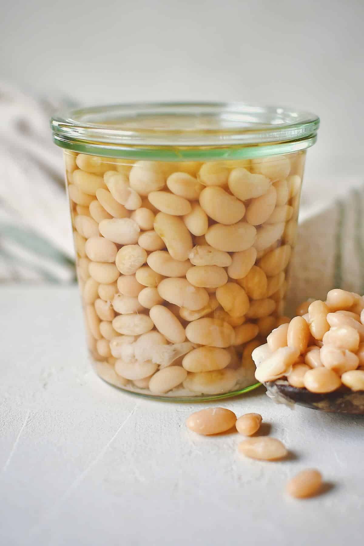 Cooked beans, stored in a glass jar for later use. some laying in a spoon on the table on the side of the jar.
