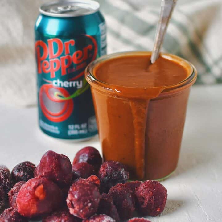 Finished Dr. Pepper BBQ Sauce in a jar that has a spoon in it, with a can of cherry dr. pepper behind it and some frozen cherries in front.