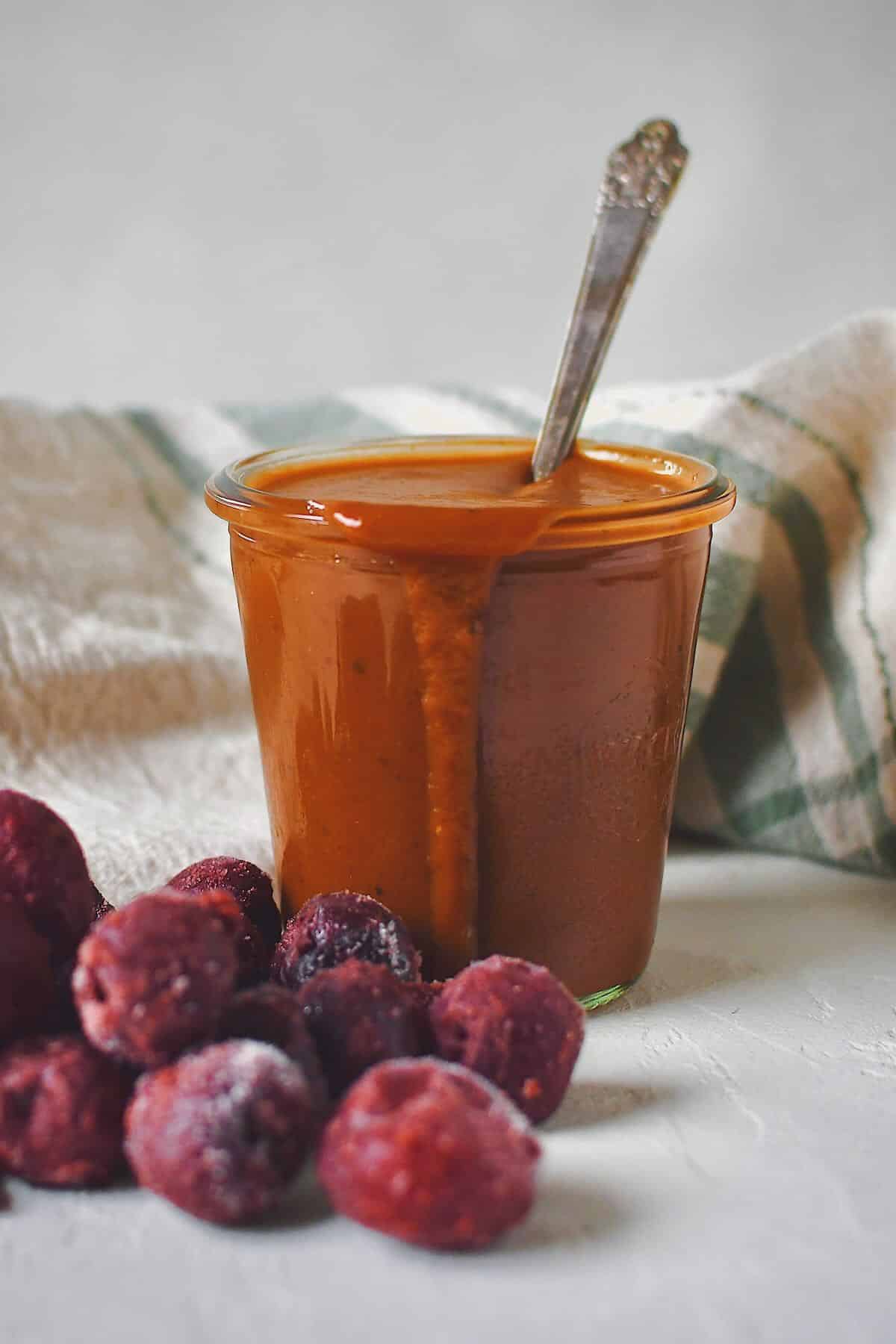 Finished Dr. Pepper BBQ Sauce in a jar that has a spoon in it, with some frozen cherries in front of it.