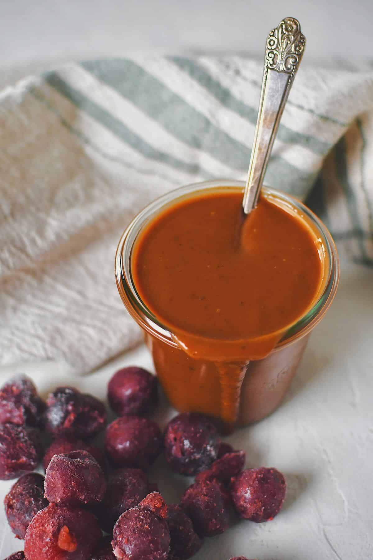 Finished Dr. Pepper BBQ Sauce in a jar that has a spoon in it, with some frozen cherries in front of it.