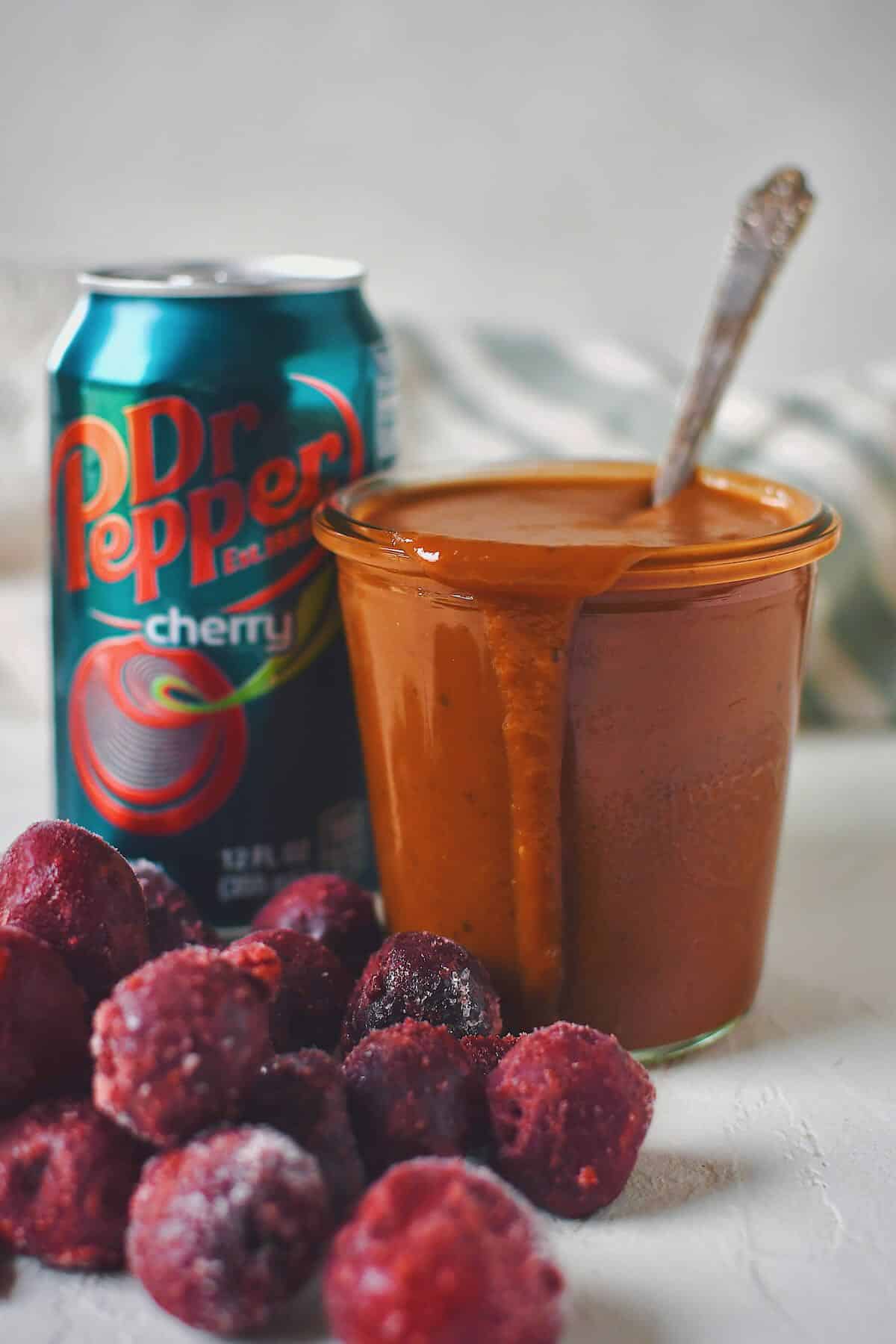 Finished Dr. Pepper BBQ Sauce in a jar that has a spoon in it, with a can of cherry dr. pepper behind it and some frozen cherries in front.