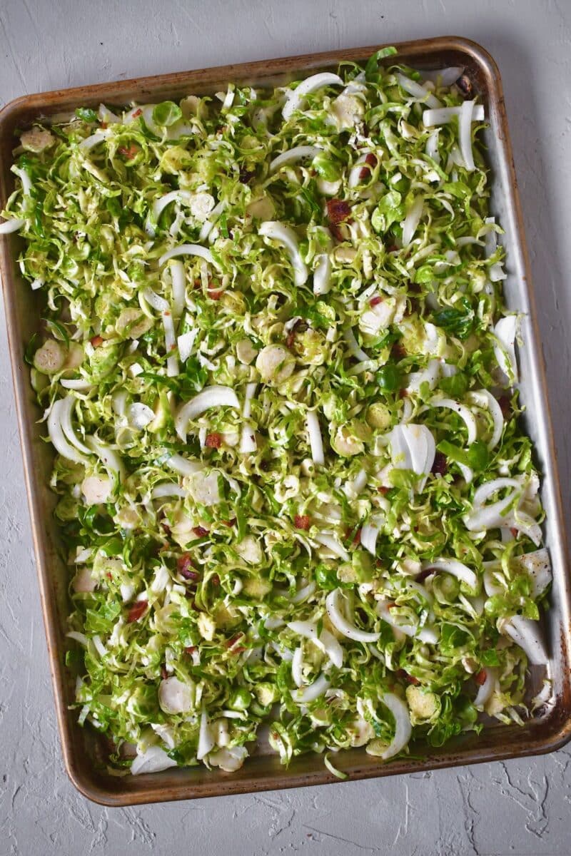 Shaved Brussel Sprouts on a baking sheet seasoned with the oil, bacon fat, salt, and pepper, and tossed with onion slices ready to be cooked.