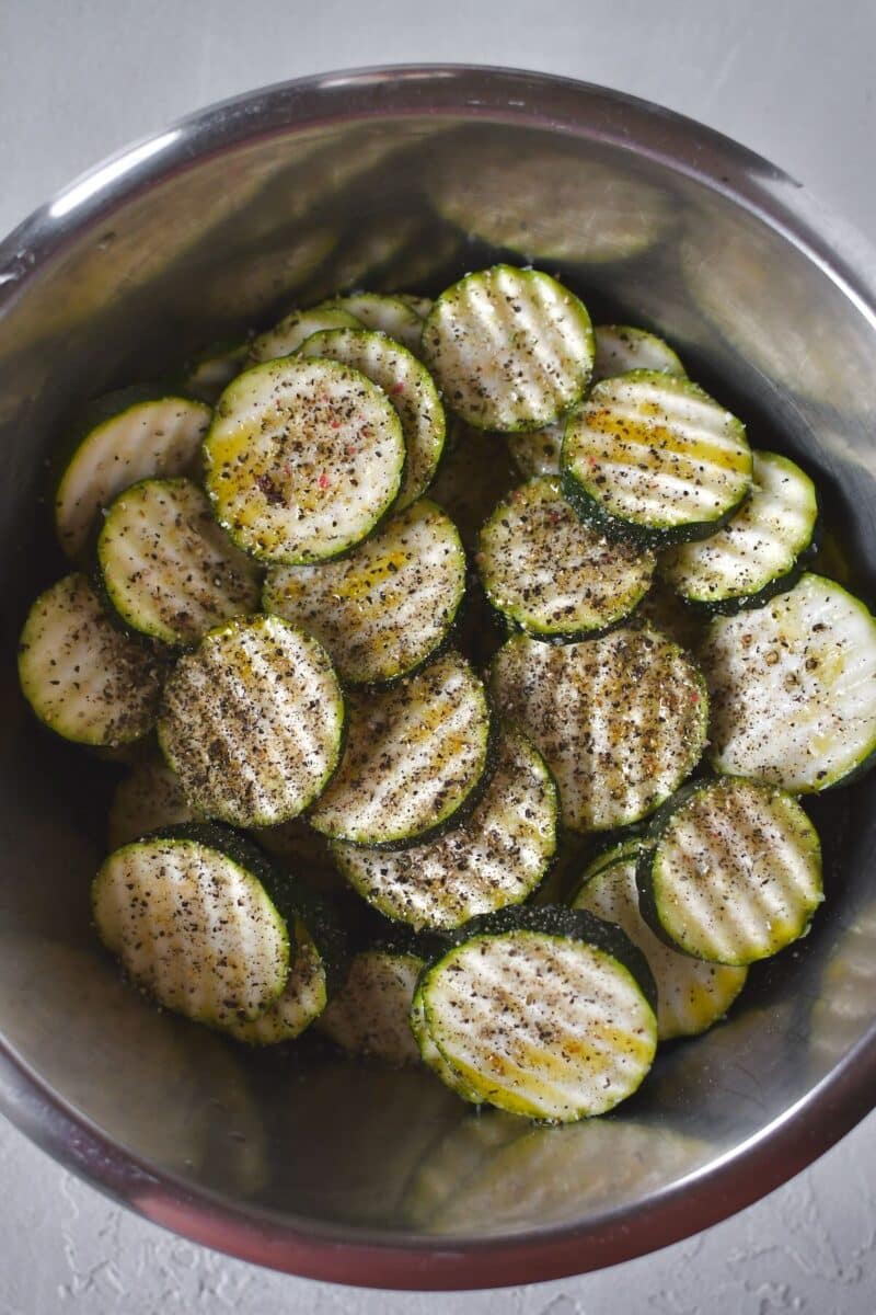 Sliced zucchini in a bowl, with olive oil drizzled over and seasoned with salt and pepper.