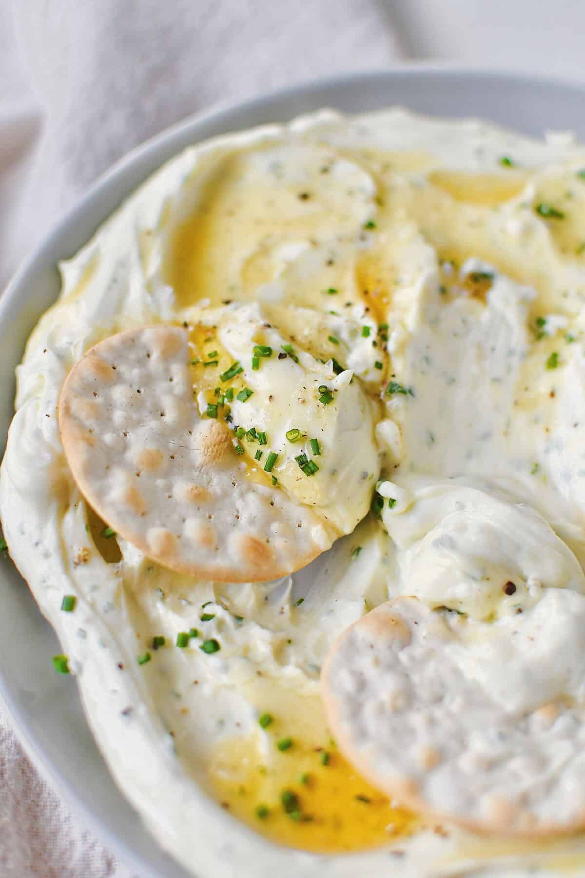 Whipped Goat Cheese spread on a plate and topped with lavender honey, chives, and sea salt. Cracker being dipped in.