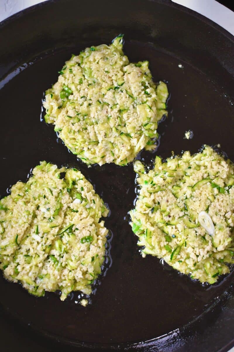 Flattened scoops of zucchini batter frying in a large pan with some oil.