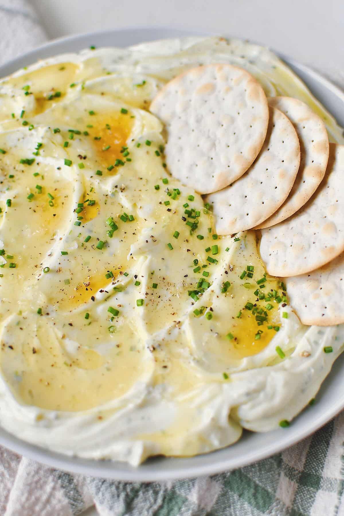 Whipped Goat Cheese spread on a plate and topped with lavender honey, chives, and sea salt. Crackers laid on top for dipping.