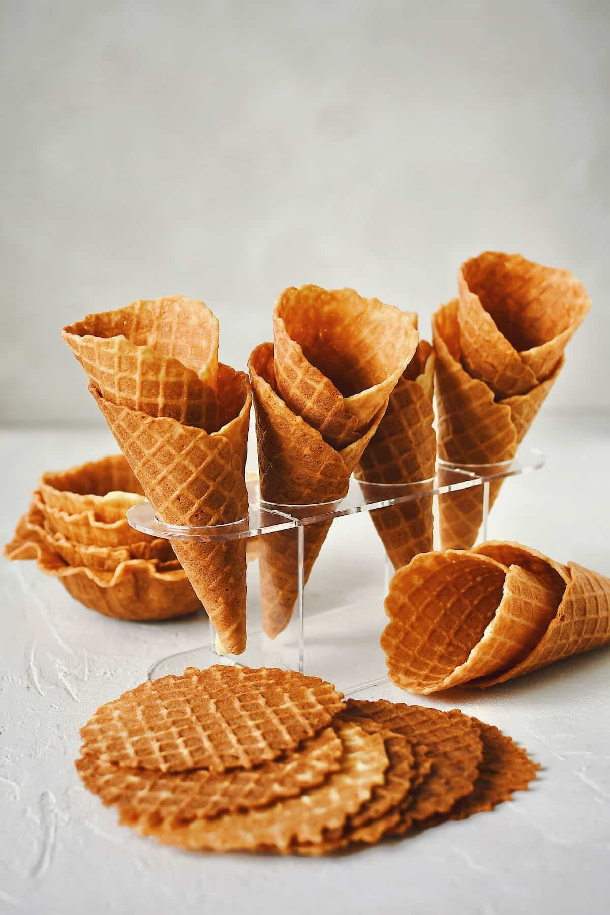 Finished Waffle Cone Recipe resting in a stand and ready to be filled with ice cream. Along with waffle bowls and flat waffle chips around it.