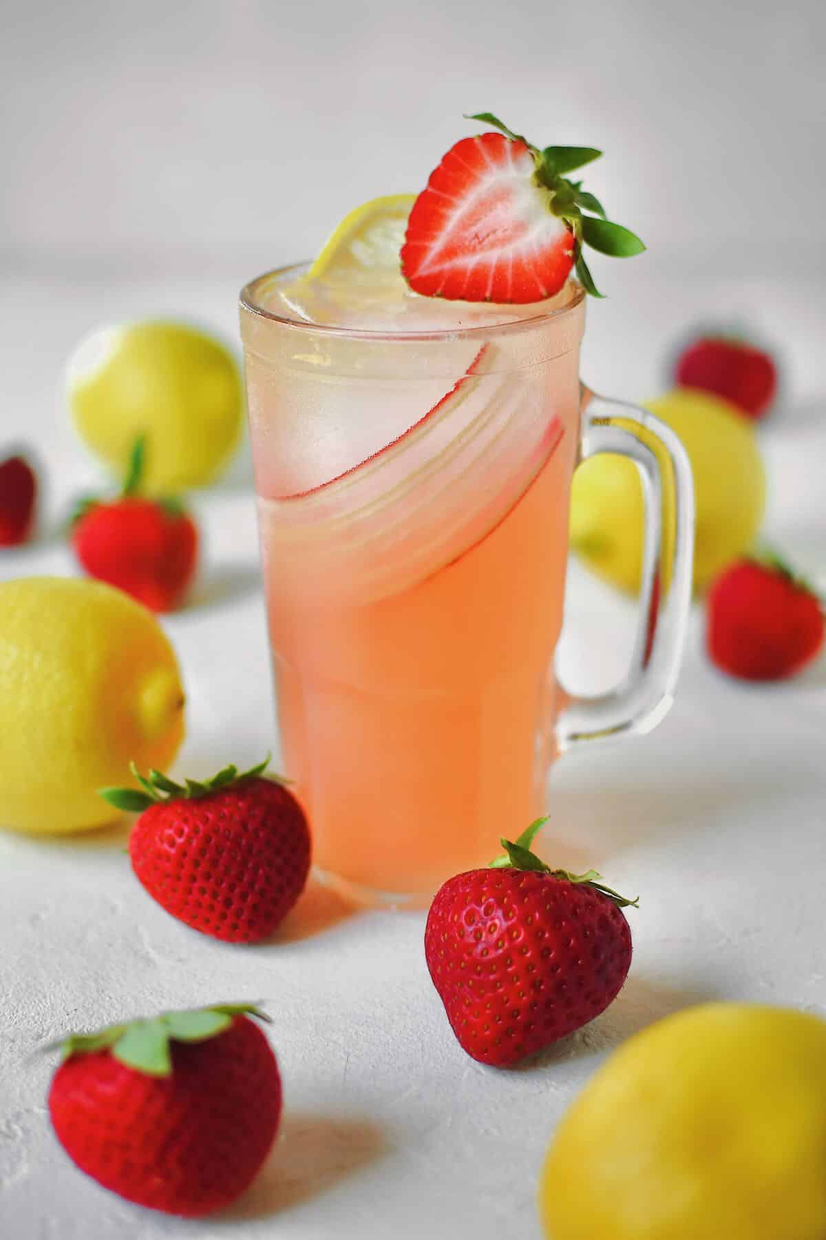 A glass of strawberry lemonade in a table with fresh strawberries and lemons around it.