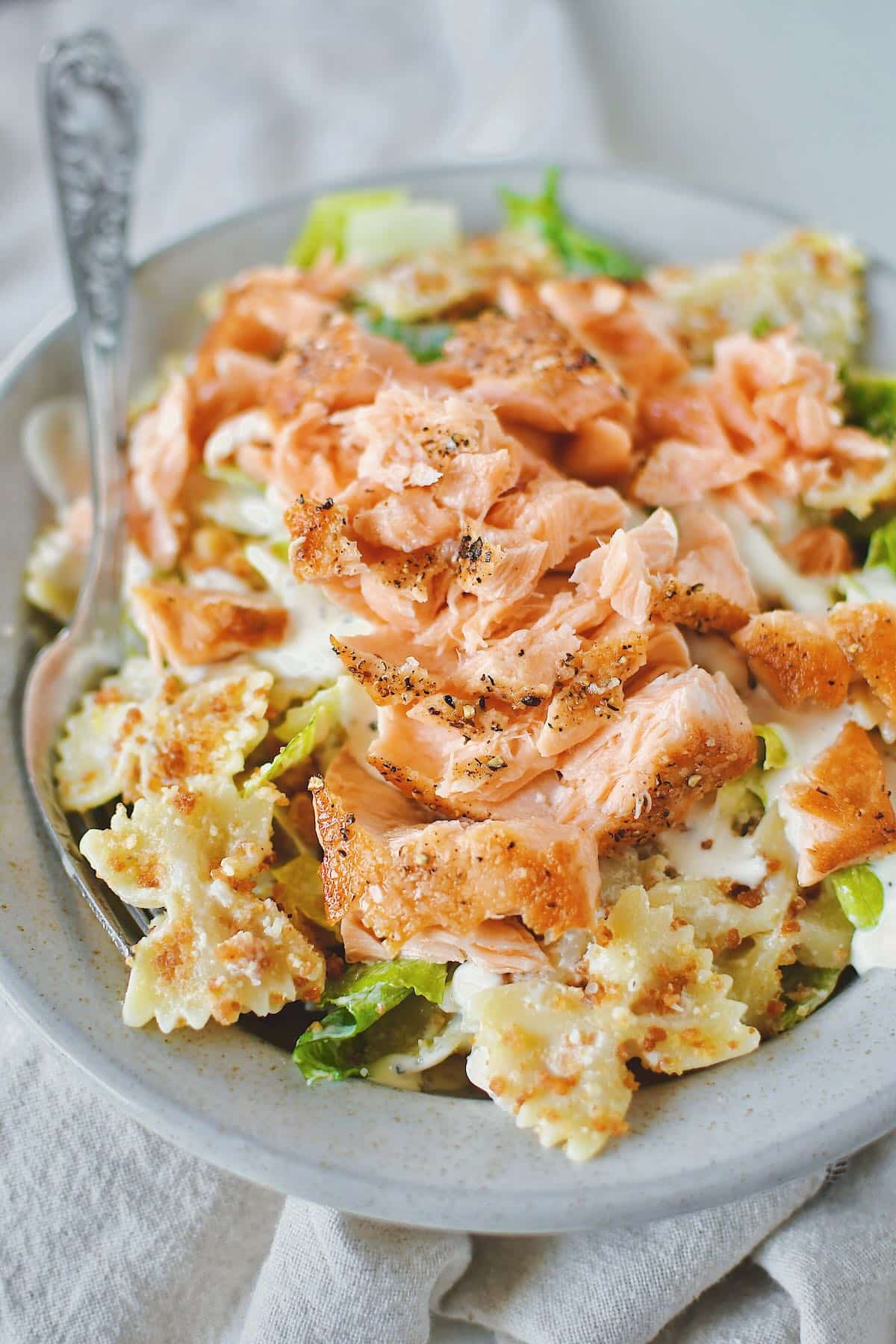 Caesar Pasta Salad served in a bowl and topped with a pan seared salmon fillet that has been cooked and flaked over the top.