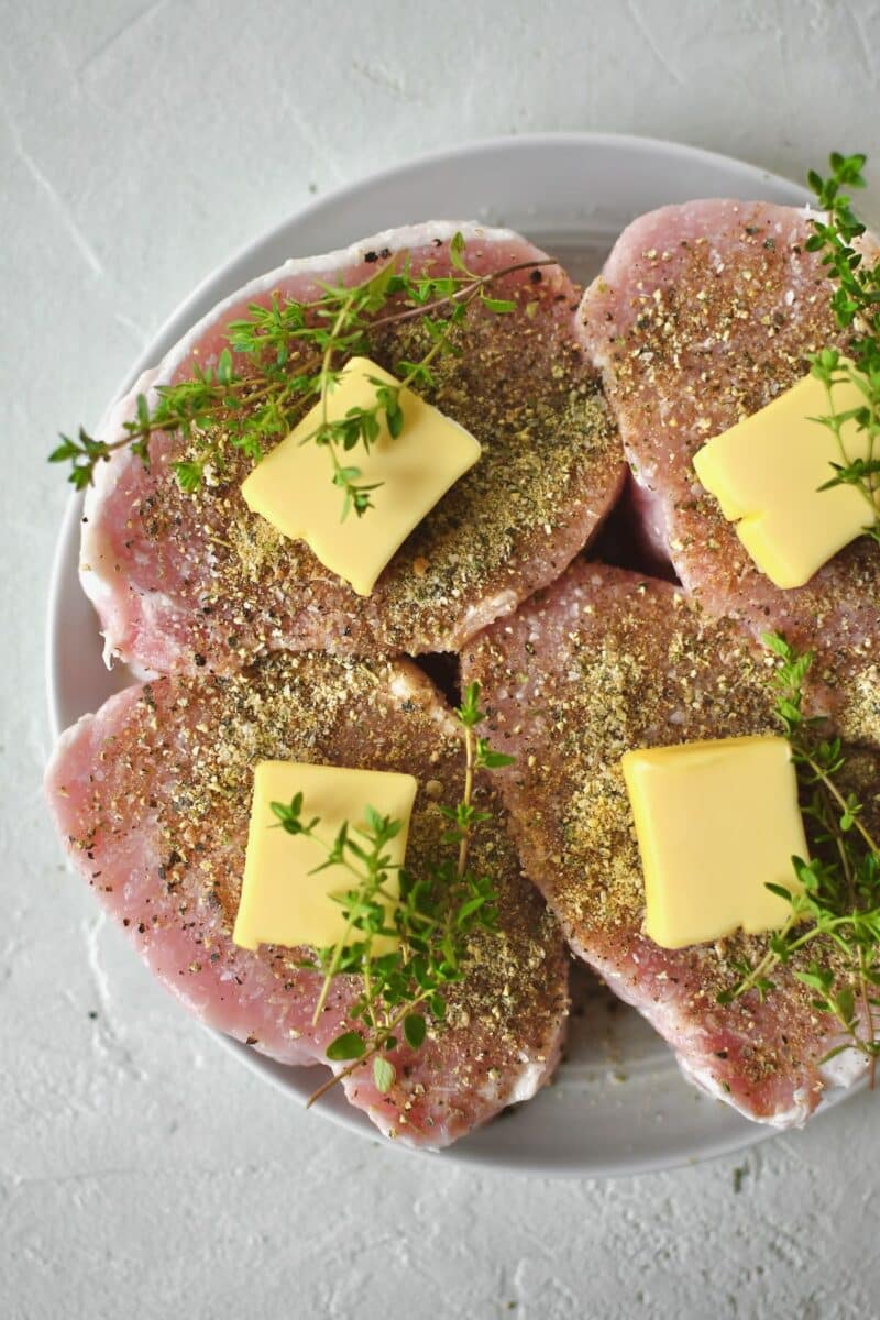 Pork Chops seasoned on both sides with salt and pepper and steak seasoning and resting on a plate. Topped with butter and thyme sprigs.