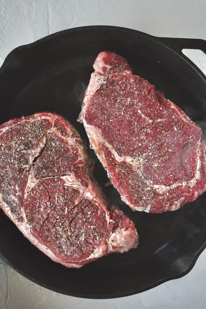 Seasoned ribeye steaks placed in a dry, hot, cast iron pan, cooking till a hard sear is achieved.