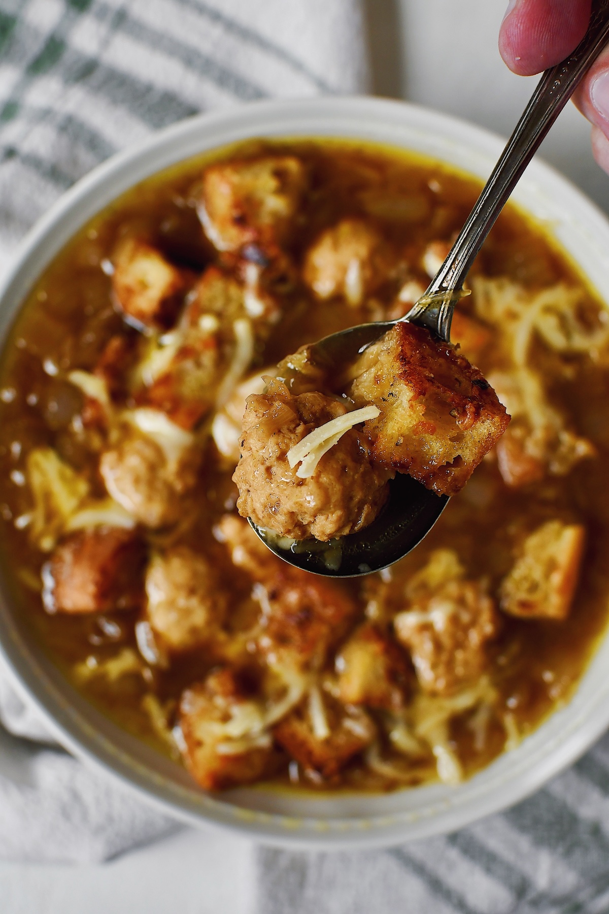 A spoon lifting a meatballs, crouton, and cheese from a bowl of Cheesy French Onion Soup.